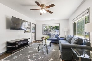 A modern living room with a ceiling fan, a flat-screen TV, a sofa, a coffee table, and a window with a view of trees. - Charming 3 Bedroom 2 Bath Home in Chico’s Peaceful Avenues (Chico)