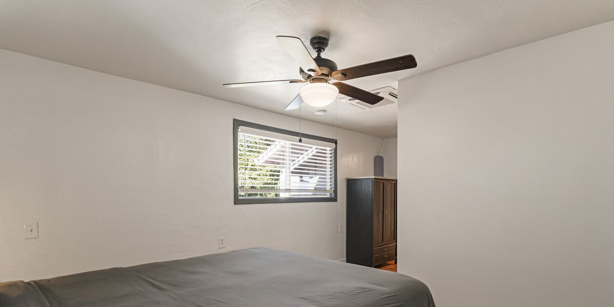 Ein Zimmer mit einem Bett, einem Deckenventilator, einem Fenster mit Jalousien und einem Holzschrank.