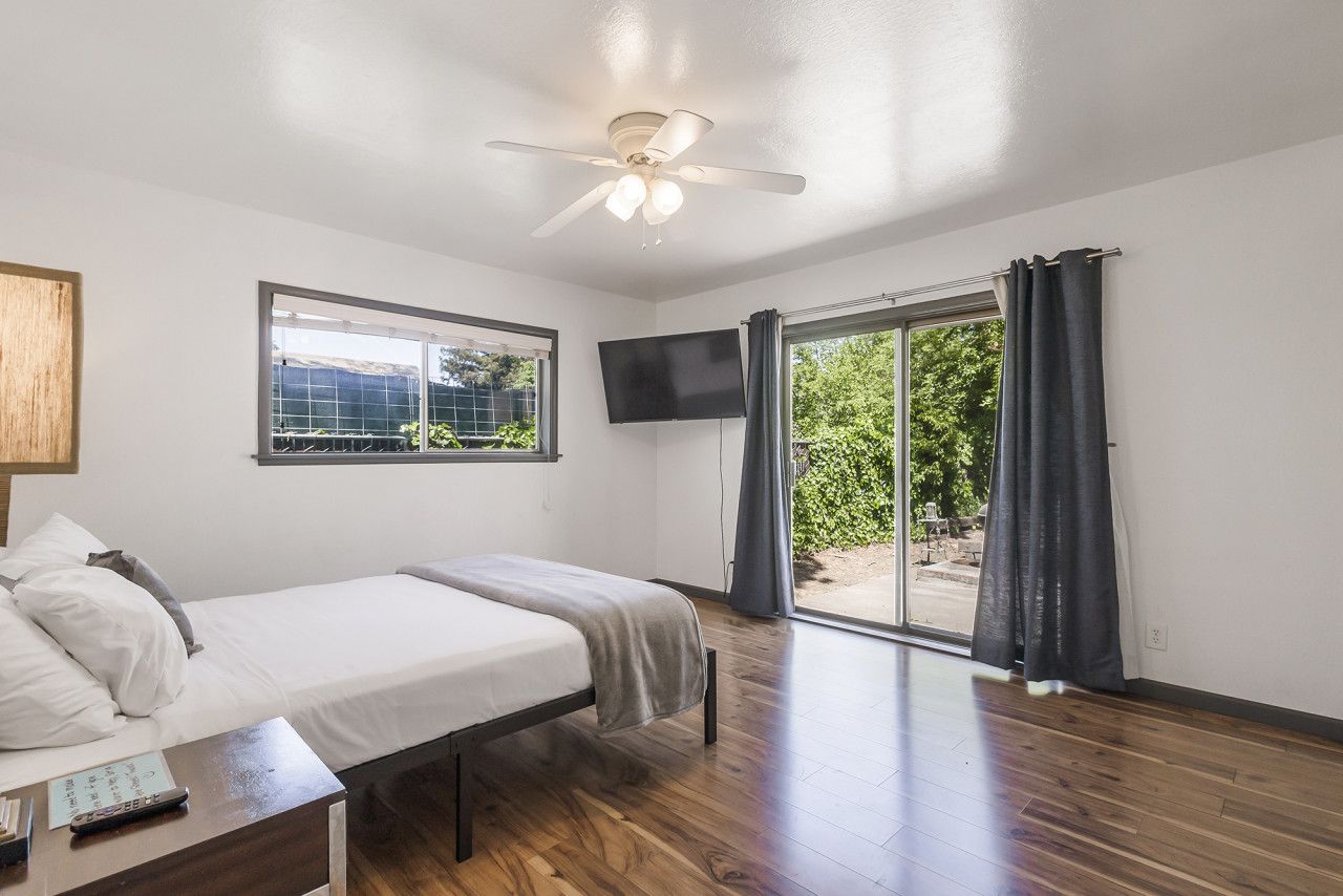 Ein Schlafzimmer mit Bett, Schreibtisch mit Stuhl, Deckenventilator, an der Wand montiertem Fernseher und einem Fenster mit Blick ins Grüne.