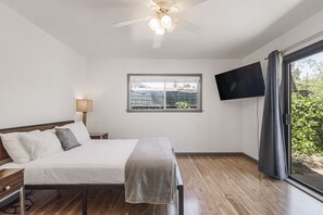 Ein Schlafzimmer mit Bett, Deckenventilator, Fenster und Fernseher, mit Blick auf Grünfläche.