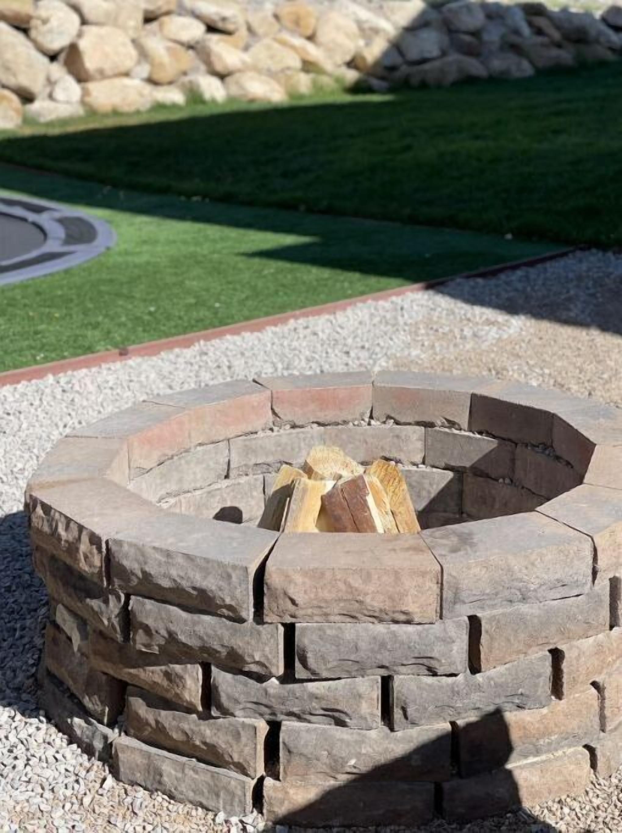 A stone fire pit with logs inside, surrounded by gravel and a grassy area with a stone wall in the background.