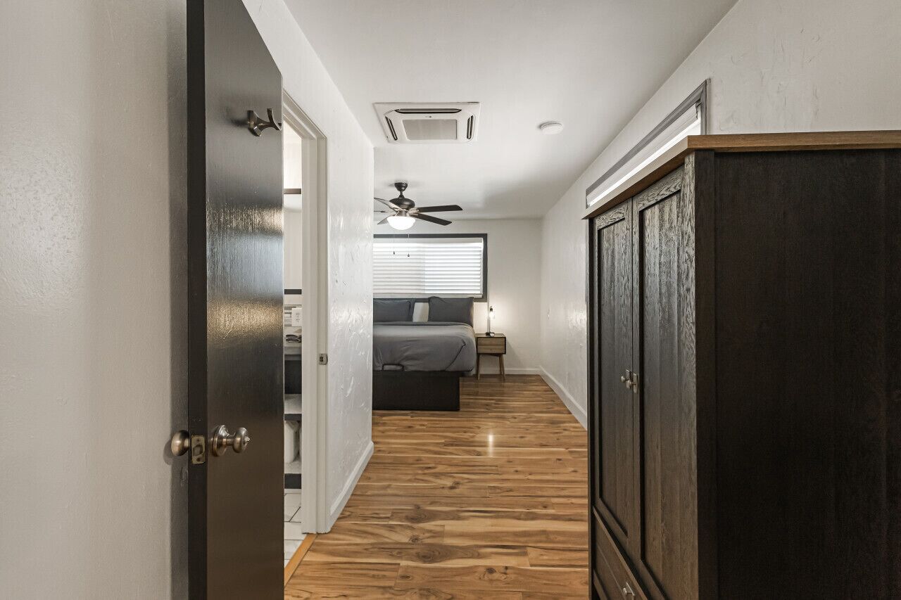 A narrow hallway with wooden flooring leading to a bedroom with a bed, bedside table, and a ceiling fan.