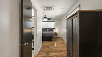 A narrow hallway with wooden flooring leading to a bedroom with a bed, bedside table, and a ceiling fan.