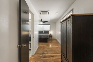 A narrow hallway with wooden flooring leading to a bedroom with a bed, bedside table, and a ceiling fan. - Charming Avenue Guest House (Chico)