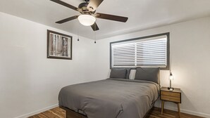 A bedroom with a bed, a ceiling fan, a bedside table with a lamp, a window with blinds, and a framed picture on the wall. - Charming Avenue Guest House (Chico)