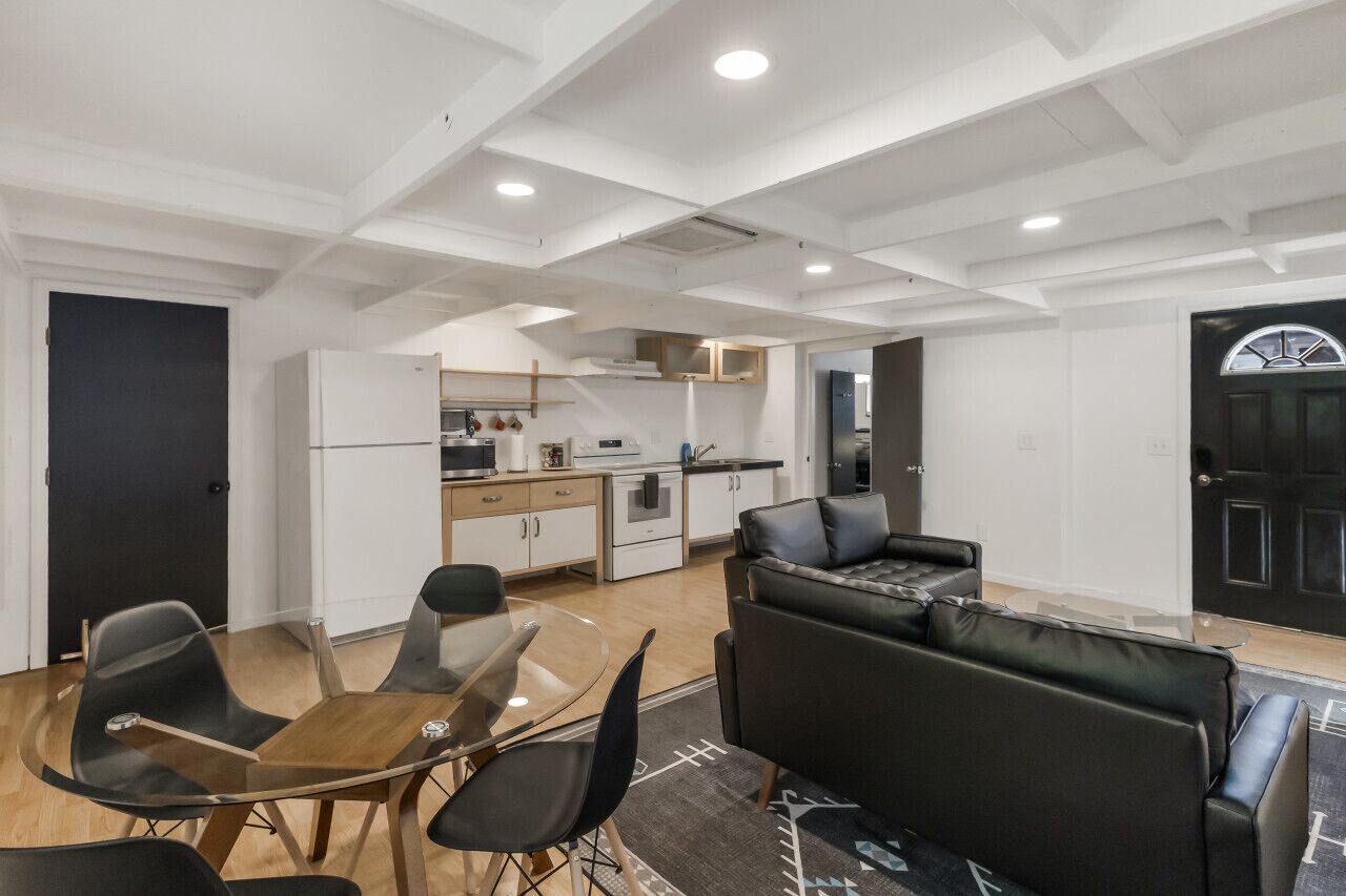 A modern kitchen with white cabinets, a dining area with a glass table and black chairs, and a black leather sofa.