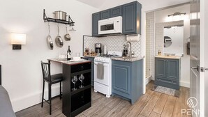 A compact kitchen with a stove, microwave, and hanging pots. There is a small dining area with a chair and wine glasses. - Chic Guesthouse Near Downtown | Stylish, Private & Cozy (Chico)
