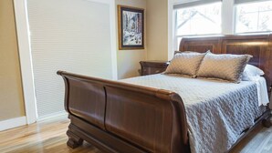 Een slaapkamer met een houten bed, beige wanden, een raam met zonwering en een ingelijste foto aan de muur.