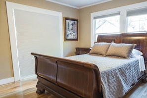 Un dormitorio con cama de madera, paredes color beige, una ventana con persianas y un cuadro enmarcado en la pared.