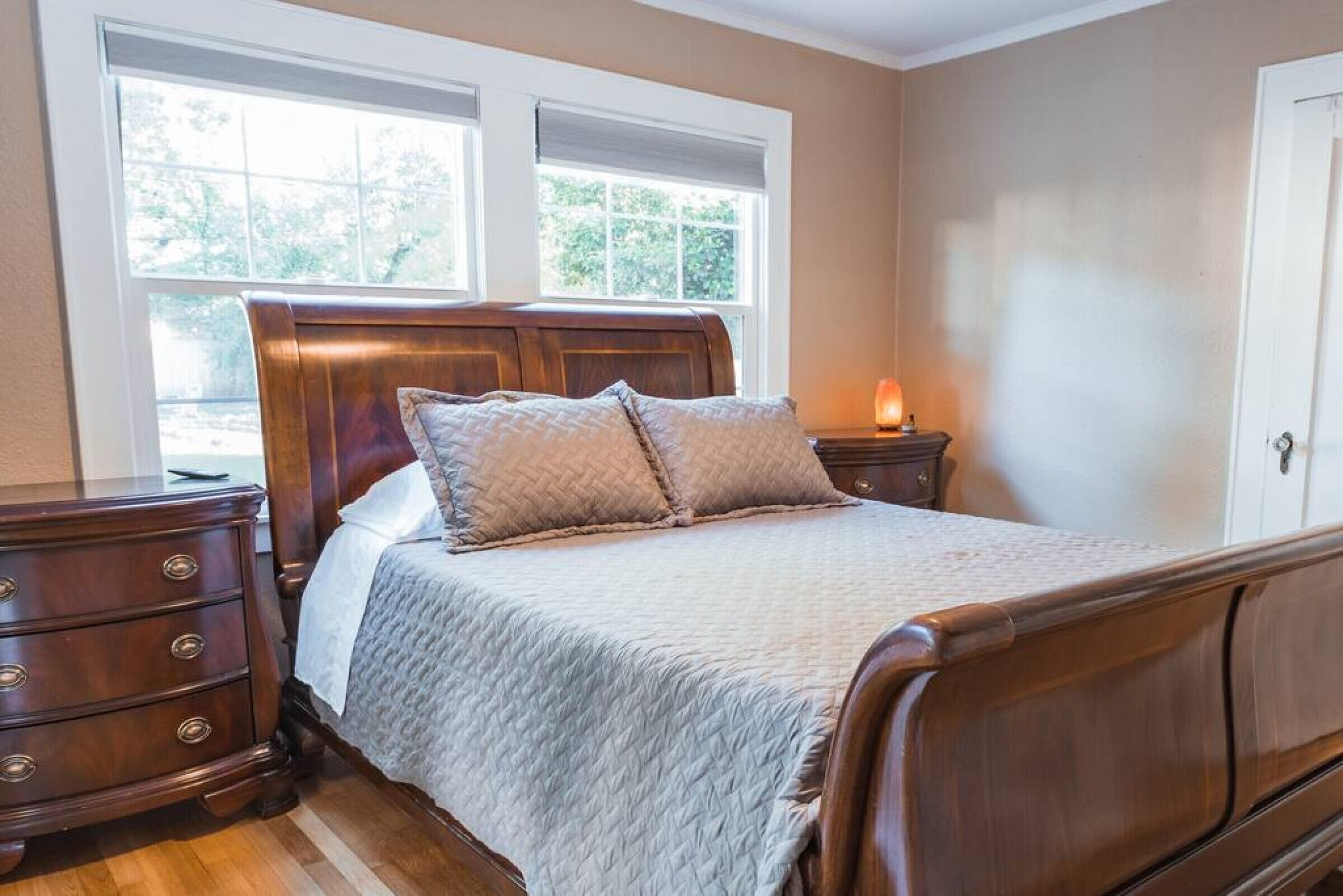 A bedroom with a wooden bed, a nightstand, a dresser, and two windows with blinds.