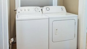 Een kleine wasruimte met een wasmachine en droger, een raam met jaloezieën, en een houten vloer.