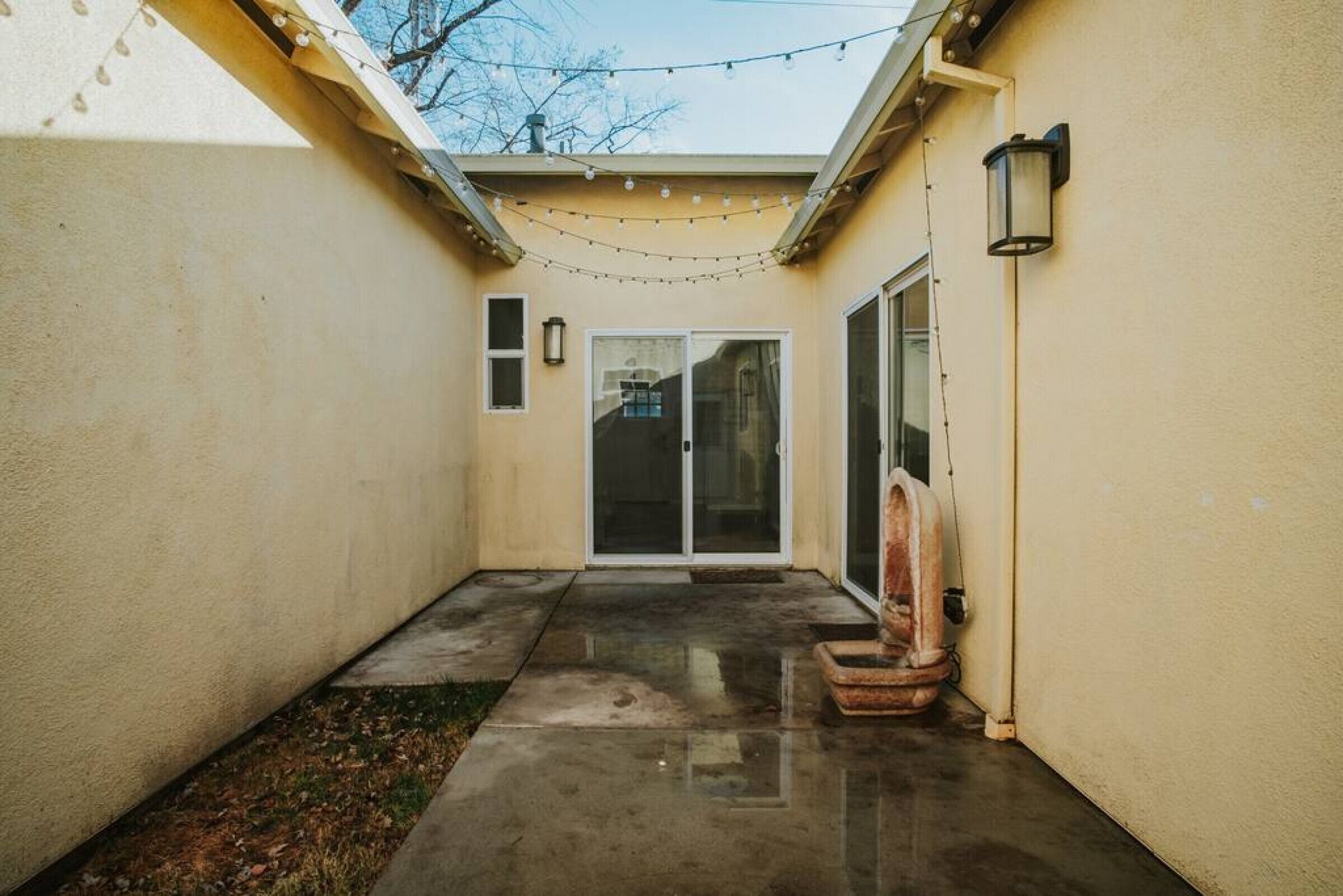 Un étroit couloir extérieur doté d’un sol en béton, d’un petit espace aménagé en jardin et d’une porte-fenêtre coulissante donnant sur un patio couvert.