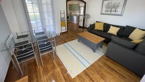 A living room with a sofa, a coffee table, a glass table with chairs, a wooden floor, and a kitchen in the background. - Gite Jeanroy a Cheerful 2 Bedroom Rural Gite (Saint-Martial-Viveyrol)