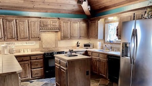A kitchen with wooden cabinets, a central island, a stainless steel refrigerator, and a window with a view. - Mountain Cabin with Multi-level Decks, Views, Hot Tub, Family Memories Made Here (Angel Fire)