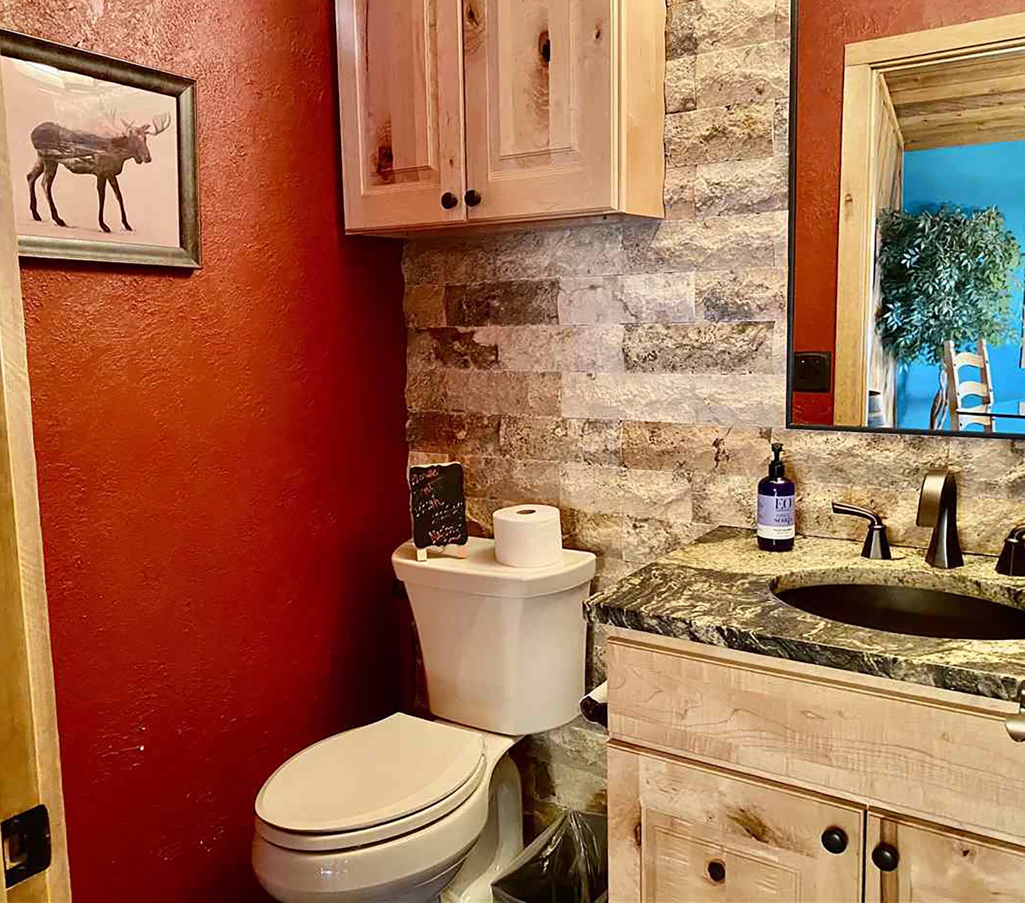 Une salle de bain avec un mur rouge, des toilettes blanches, un lavabo avec un plan de travail en marbre et des meubles en bois.