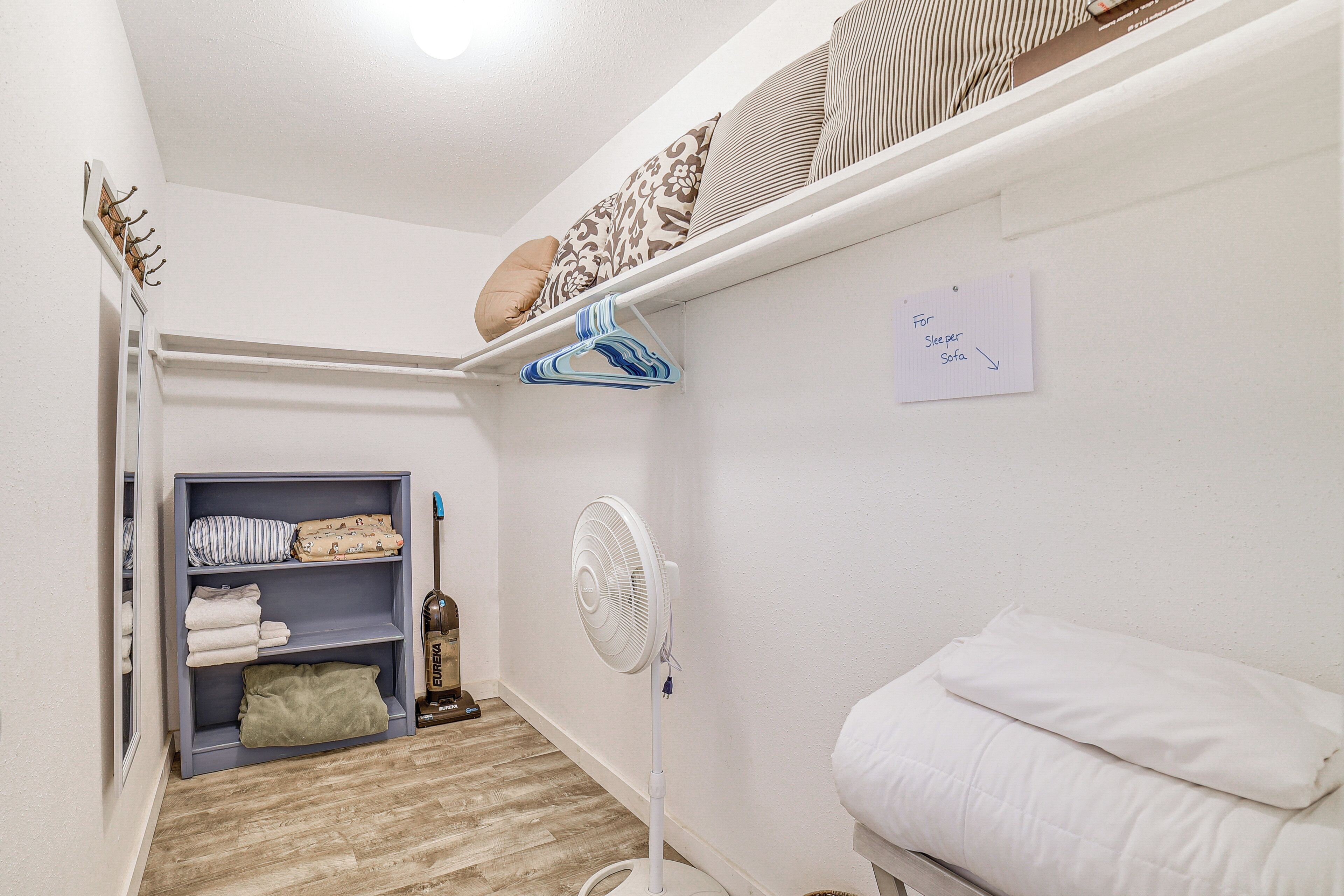 Ein schmaler Flur mit weißer Wand, einem Regal mit gefalteten Decken, einem Standventilator und einem Zettel an der Wand.