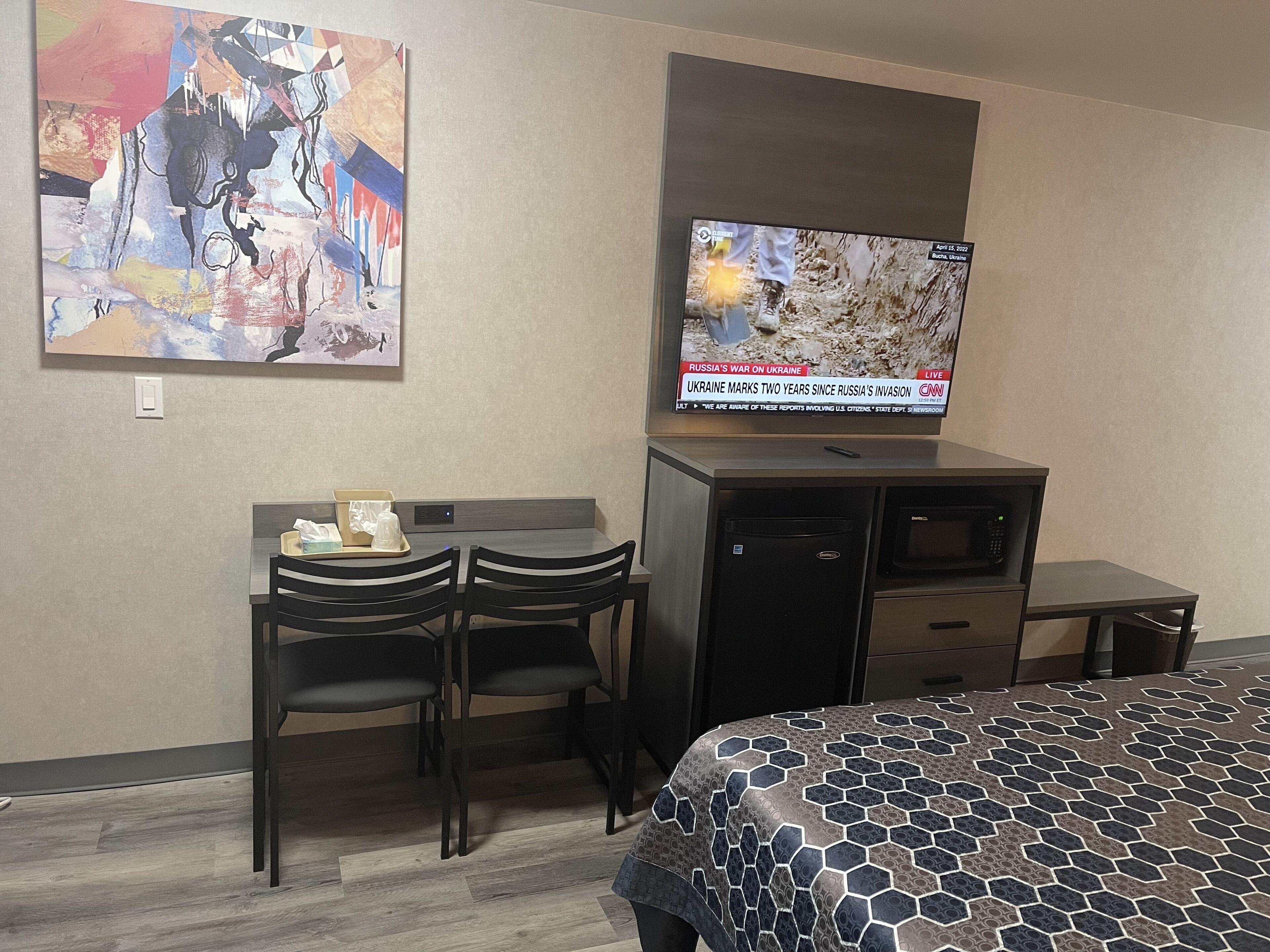 Habitación de hotel con cama, escritorio con dos sillas, televisor montado en la pared y un cuadro en la pared.