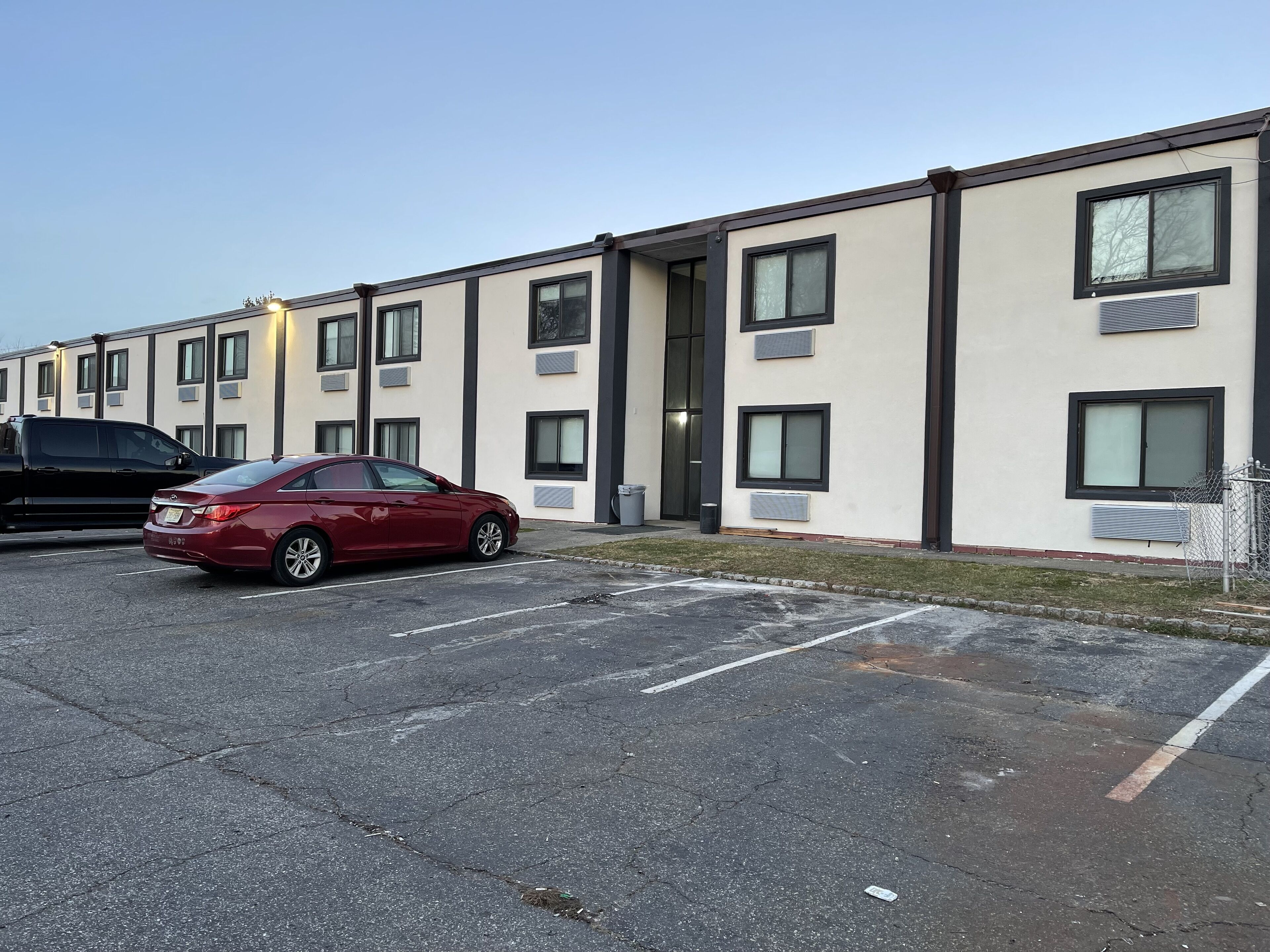 A parking lot with a red car and a building with multiple windows and air conditioning units.