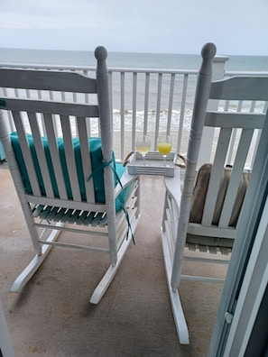 Un balcon donnant sur la plage, avec un fauteuil à bascule blanc et une petite table sur laquelle sont posés deux verres remplis de boisson.