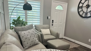 A living room with a beige sectional sofa, patterned pillows, a small ottoman, a floor lamp, and a large wall clock. - Country Living in the City (Mesa)
