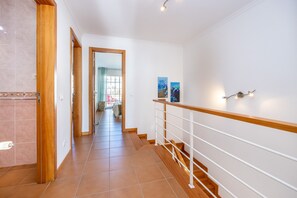 Een hal met een houten vloer, witte wanden en een witte leuning. Twee schilderijen hangen aan de muur en een deur leidt naar een andere kamer.