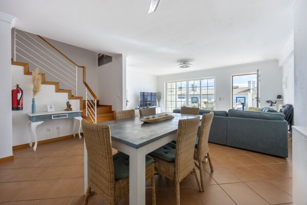 A spacious living area with a dining table, wicker chairs, a sofa, and a staircase.
