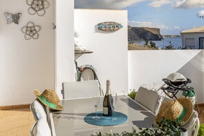 Un patio con una mesa lista para dos personas, una botella de vino y una copa. Hay dos sombreros tejidos sobre la mesa, un casco y un scooter.