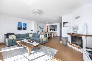 Een moderne woonkamer met een bank, salontafel en open haard. Er is een eethoek met een tafel en stoelen. De kamer heeft witte wanden en betegelde vloeren.