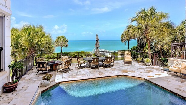 A pool area with a view of the ocean, outdoor furniture, and palm trees.