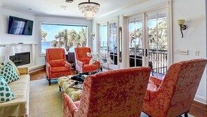 A living room with a fireplace, a television mounted on the wall, and a large window overlooking the ocean.