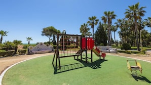 Un parque infantil con un tobogán y estructuras para escalar, rodeado de palmeras y un cielo despejado.