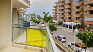 Un balcón con piso amarillo, que da a una calle con coches y un edificio con varios balcones.