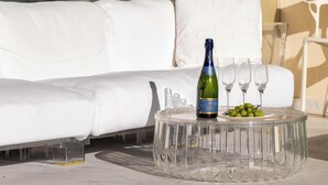 Ein beigestellter Tisch aus Glas mit einer Champagnerflasche und einer Weintraubentafel, auf einem Balkon mit einem weißen Sofa und einem weiteren Glastisch im Hintergrund.