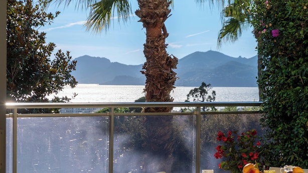 Ein Zimmer mit großem Fenster, das Blick auf ein Gewässer und Berge, eine Palme, einen Tisch mit Früchten und eine Blumenvase bietet.