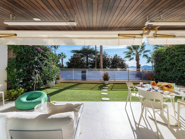 A modern outdoor patio with a dining table set for two, surrounded by lush greenery and a clear view of the ocean.