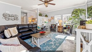 A living room with a black leather sofa, a coffee table, and a blue star-patterned rug. There is a ceiling fan, a white wall with decorative items, and a large window with a view of the outdoors.