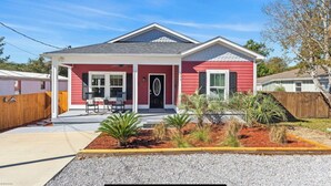 A single-story house with a red exterior, a front porch with two chairs, and a small garden area with various plants.