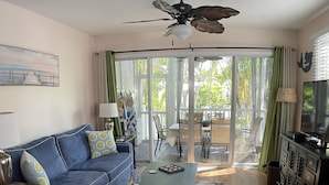 A living room with a blue sofa, a coffee table, a ceiling fan, and a view through a glass door to an outdoor area. - Poolside Paradise in Key West Golf Club (Key West)