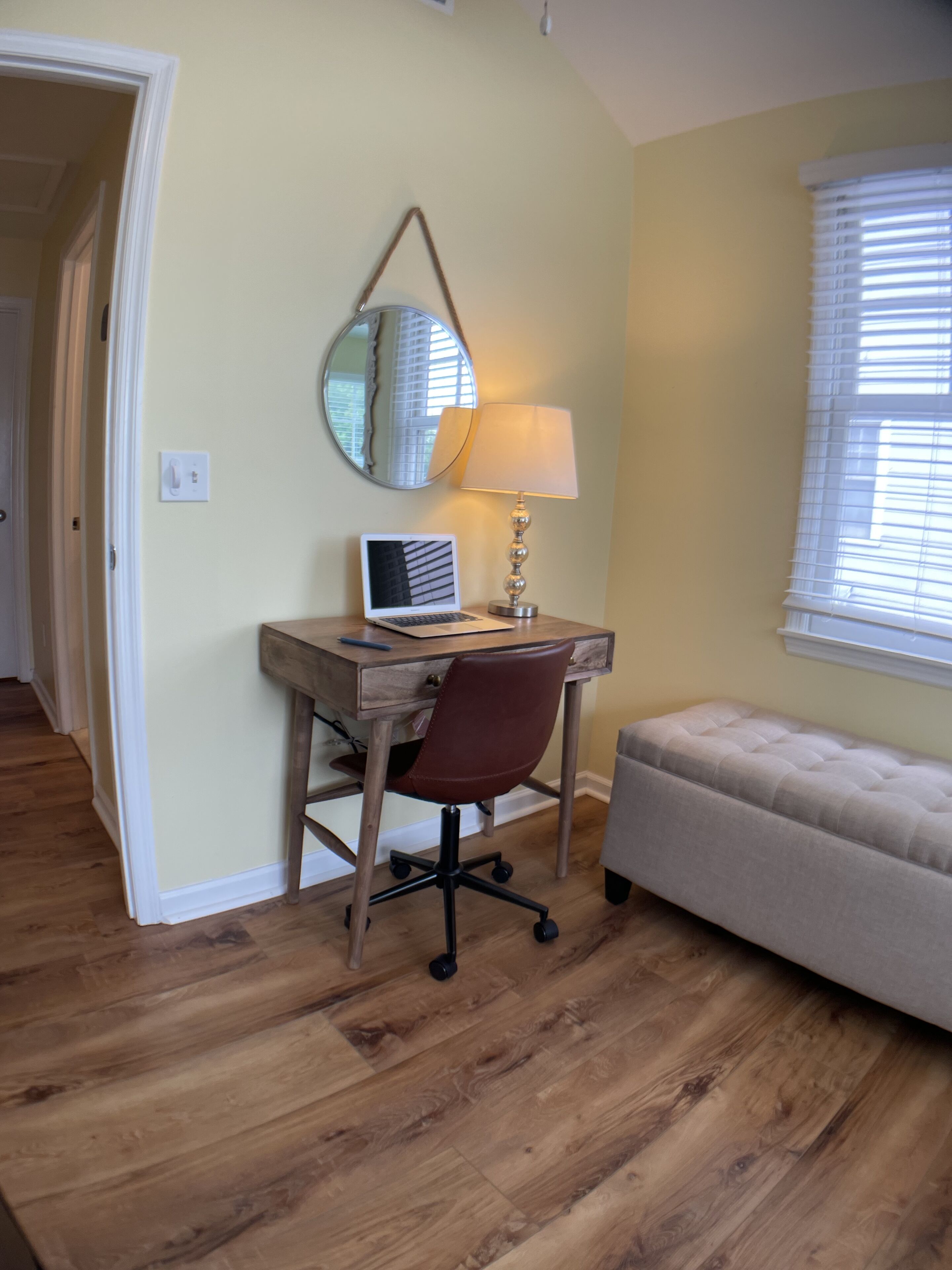 Una habitación con un escritorio de madera, una laptop, una silla, una lámpara, un espejo, una ventana con persianas y un sofá.