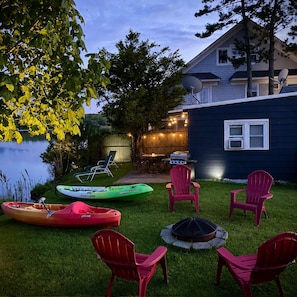 Un patio trasero con kayaks, sillas, un pozo de fuego y una zona de comedor con luces colgantes.