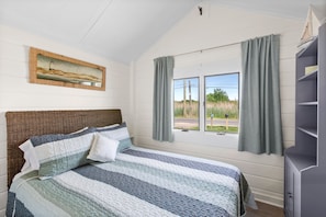 A bedroom with a bed, a window with curtains, a bookshelf, and a painting on the wall. - Cozy cottage on Fort Pond Lake. (Montauk)
