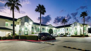 A multi-story hotel with a parking lot, palm trees, and a clear sky. - Palace Inn I10East & John Ralston (Houston)