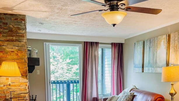 A living room with a brown leather sofa, a ceiling fan, a stone wall, a window with curtains, and a view of greenery outside. - Blue Knob Big Snow Condo  (Claysburg)