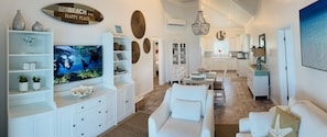 Uma sala de estar moderna com televisão de tela plana, móveis brancos, arte decorativa nas paredes e vista para a cozinha.