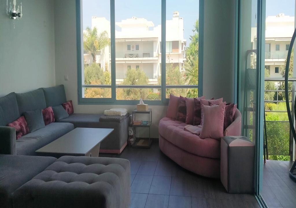 A modern living room with a grey sectional sofa, a pink sofa, and a grey ottoman. There is a small table and a side table with a lamp. Large windows provide a view of buildings and greenery outside.