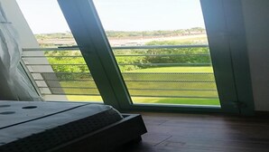 Un dormitorio con cama, ventana y vista a un paisaje verde, además de barandilla metálica.