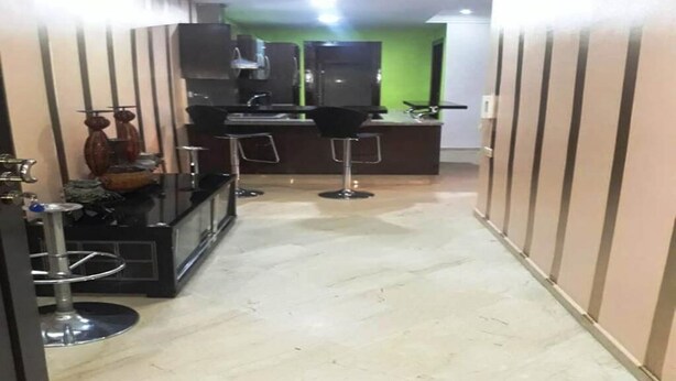 Un pasillo estrecho con una zona de kitchenette equipada con refrigerador, microondas y cafetera. Hay dos taburetes de bar y una pequeña mesa con adornos. Las paredes están revestidas de paneles, y el piso está cubierto de baldosas.