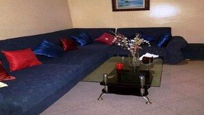 Un salon avec un canapé bleu foncé, des coussins rouges et bleus, une table basse en verre ornée d’un vase de fleurs et un tableau encadré accroché au mur.