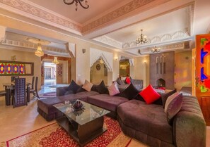 Un salon spacieux avec un grand canapé d’angle, une table basse en verre et un tapis rouge à motifs. On aperçoit un lustre et une arche décorative au fond.