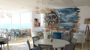 A dining area with round tables and chairs, a large mural on the wall, and a view of the ocean through large windows. - Residence La Source Taghazout 5217 (Taghazout)